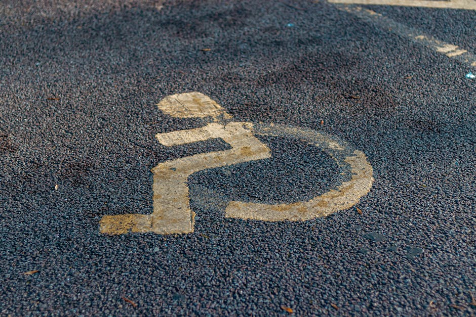 Close-up of a yellow wheelchair accessibility symbol painted on a rough-textured asphalt surface outside a property, indicating the area is designated for disabled access or parking. The symbol shows a simplified figure seated in a wheelchair with a circular wheel and an outline of legs and arms, positioned on the pavement near a home or building. The surrounding asphalt has small stones and debris, with the paint slightly worn in some areas. This image is relevant in the context of house removals and moving logistics, highlighting considerations such as parking permits or access requirements during furniture transport and loading processes, which [COMPANY_NAME], such as Man With a Van Anerley, manages as part of their residential removal services.