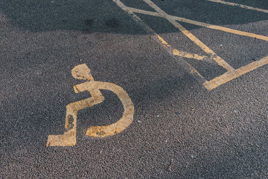 A close-up view of a disabled parking space marked by a worn, yellow wheelchair symbol painted on dark asphalt paving. The painted symbol shows a person seated in a wheelchair with a circular wheel at the base, positioned within a designated parking bay with faded yellow lines indicating parking boundaries. The surrounding surface is textured with small pebbles embedded in the asphalt. Although no vehicles or furniture are visible, this scene reflects parking regulations related to home relocation or moving services. The image is captured in natural lighting, highlighting the worn condition of the parking markings, which may impact access for moving or transportation logistics. Man With a Van Anerley offers local removals and furniture transport, supporting moves that require careful parking management and vehicle access planning, especially in areas with narrow streets or restricted parking zones.