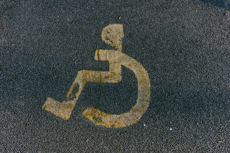 Close-up of a yellow wheelchair accessibility symbol painted on a rough-textured asphalt surface outside a property, indicating the area is designated for disabled access or parking. The symbol shows a simplified figure seated in a wheelchair with a circular wheel and an outline of legs and arms, positioned on the pavement near a home or building. The surrounding asphalt has small stones and debris, with the paint slightly worn in some areas. This image is relevant in the context of house removals and moving logistics, highlighting considerations such as parking permits or access requirements during furniture transport and loading processes, which [COMPANY_NAME], such as Man With a Van Anerley, manages as part of their residential removal services.