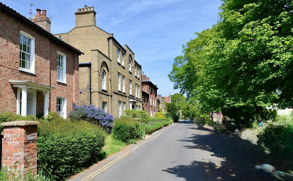 A residential street scene featuring a row of traditional brick and stone houses with white-framed windows and small front gardens, some with flowering shrubs and greenery. The street is paved and qui