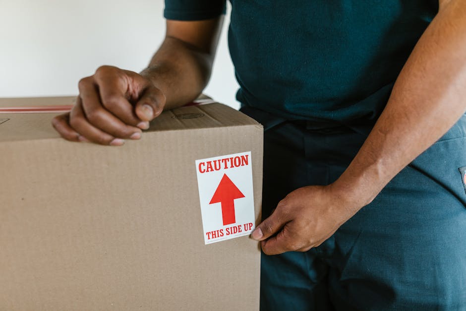 Close-up of a person in dark clothing holding a large cardboard moving box with both hands, applying a red and white 'Caution' label that reads 'This Side Up' with an upward-pointing arrow. The background shows an indoor setting with a neutral-colored wall and a wooden floor, indicating the packing process during a home relocation or furniture transport. The box appears to be carefully handled, emphasizing proper loading techniques common in professional removals services. This scene is part of a house moving or packing effort, likely assisted by [COMPANY_NAME], which offers removals and moving logistics in the Anerley area, as suggested by the context of the webpage title concerning SE20 moving tips and removals near Anerley Station.