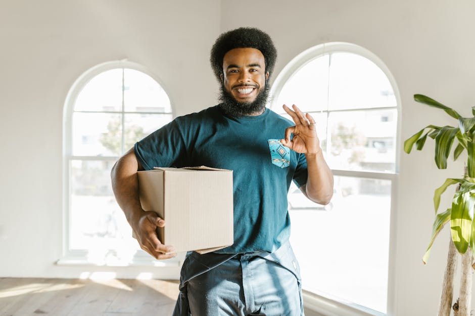 A smiling man with a curly afro hairstyle and a beard, holding a cardboard box wrapped in brown packing paper against his hip inside a bright room with large arched windows, natural light illuminating the space. He is making an 'okay' gesture with his right hand and wearing a teal T-shirt with a small pocket containing a patterned cloth. The room has white walls, and a green potted plant is visible on the right side. This image depicts the process of packing and home relocation, with the man representing a mover from Man With a Van Anerley, involved in furniture transport and loading efforts as part of a house move. The scene emphasizes the packing and moving logistics associated with professional removals near Anerley Station, showcasing the preparation stage of a house removal service.
