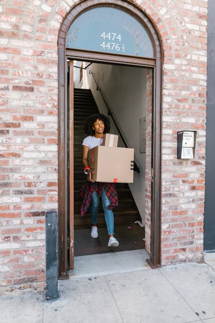 Close-up of a person in dark clothing holding a large cardboard moving box with both hands, applying a red and white 'Caution' label that reads 'This Side Up' with an upward-pointing arrow. The background shows an indoor setting with a neutral-colored wall and a wooden floor, indicating the packing process during a home relocation or furniture transport. The box appears to be carefully handled, emphasizing proper loading techniques common in professional removals services. This scene is part of a house moving or packing effort, likely assisted by [COMPANY_NAME], which offers removals and moving logistics in the Anerley area, as suggested by the context of the webpage title concerning SE20 moving tips and removals near Anerley Station.